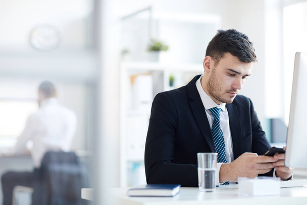 Grave giovane uomo d'affari seduto sul posto di lavoro di fronte al computer e sms in smartphone
 - Foto, immagini