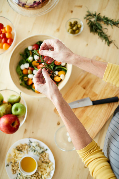 Directly above view of unrecognizable woman standing at table with dishes and adding rosemary into vegetable salad while preparing for dinner - Photo, image