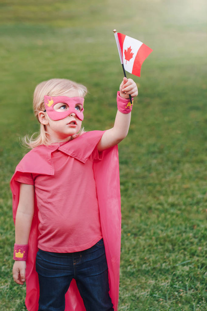 Niña caucásica en cotume de superhéroe y máscara con bandera canadiense ondeante. Chica celebrando la fiesta nacional de Canadá día en el parque de verano al aire libre. Concepto de fuerza, potencia y protección
.  - Foto, Imagen