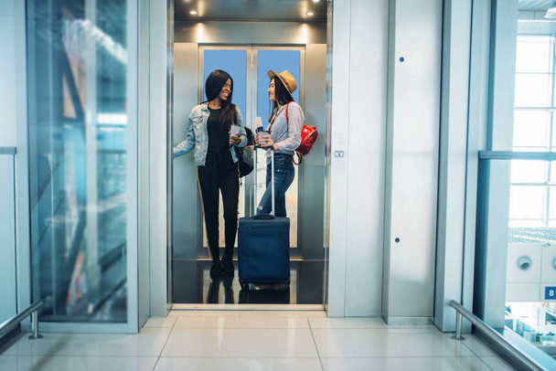 Две женщины с детьми в лифте аэропорта. Пассажиры с багажом в аэровокзале, вид сзади, счастливое путешествие белых и черных дам, летние путешествия
 - Фото, изображение