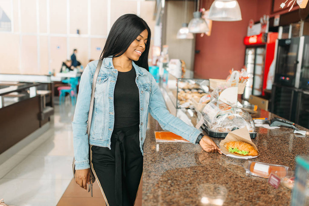 Black female traveler buying burger in airport waiting area. Passenger choosing food in air terminal, happy journey of attractive lady, summer travel on vacation - Foto, Bild