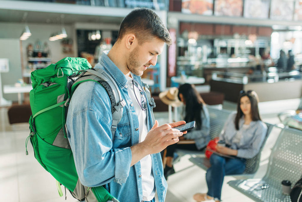 Male tourist with backpack holds mobile phone, female travelers waiting for departure in airport on background. Passengers with baggage in air terminal, happy journey, summer travel - Foto, Bild