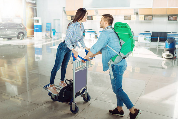 Αρσενικό τουριστικό ρολό το κάρο με τις αποσκευές και τη γυναίκα του στο αεροδρόμιο. Επιβάτες με αποσκευές στον τερματικό αεροσταθμό, ευτυχισμένο ταξίδι, καλοκαιρινές μετακινήσεις - Φωτογραφία, εικόνα