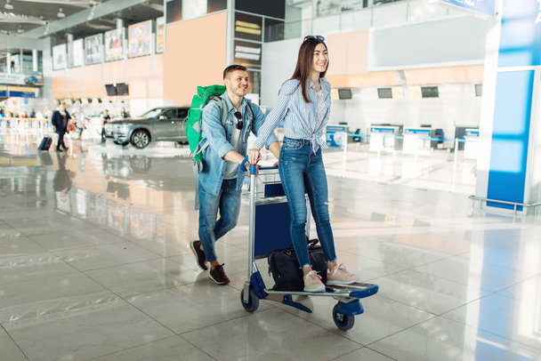 Αρσενικό τουριστικό ρολό το κάρο με τις αποσκευές και τη γυναίκα του στο αεροδρόμιο. Επιβάτες με αποσκευές στον τερματικό αεροσταθμό, ευτυχισμένο ταξίδι, καλοκαιρινές μετακινήσεις - Φωτογραφία, εικόνα