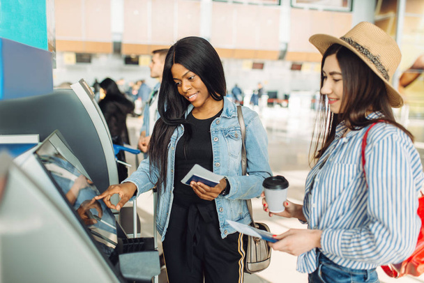 Two female travelers withdraw cash at ATM in airport. Passengers with baggage in air terminal, back view, happy journey of white and black ladies, summer travel - Foto, Imagen