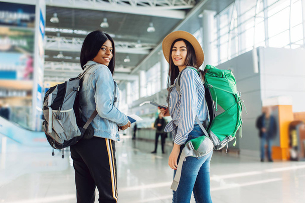 Dwie kobiece turyści z plecaki na międzynarodowym lotnisku. Pasażerowie z bagażem w terminalu lotniczym, widok z tyłu, szczęśliwa podróż białych i czarnych pań, letnia podróż szczęśliwych kobiet - Zdjęcie, obraz