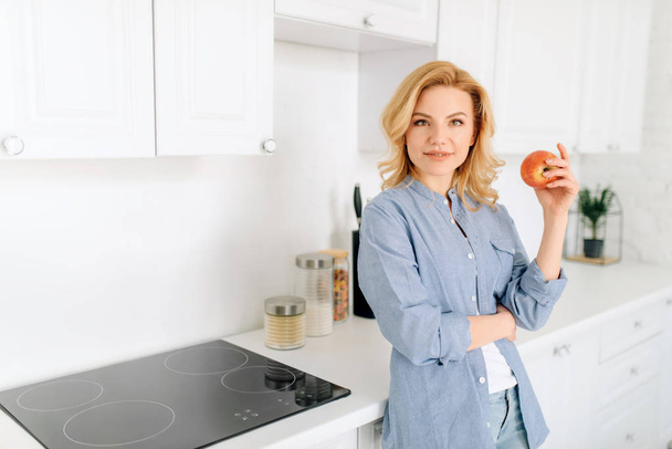 Portrait of woman with apple poses on the kitchen with snow-white interior. Female person at home in the morning, healthy nutrition and lifestyle - Zdjęcie, obraz