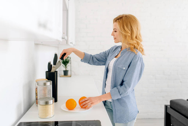 Ευτυχισμένη γυναίκα με μαχαίρι που ετοιμάζει πορτοκάλια, πρωινό στην κουζίνα. Θηλυκό άτομο στο σπίτι το πρωί, υγιεινή διατροφή και lifestyle - Φωτογραφία, εικόνα