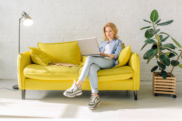 Young lady reading a book on cozy yellow couch, living room in white tones on background. Attractive female person with magazine sitting on sofa at home - Photo, image