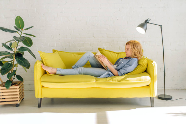 Young woman lying on cozy yellow couch and reading a book, living room in white tones on background. Attractive female person with magazine sitting on sofa at home - Fotoğraf, Görsel