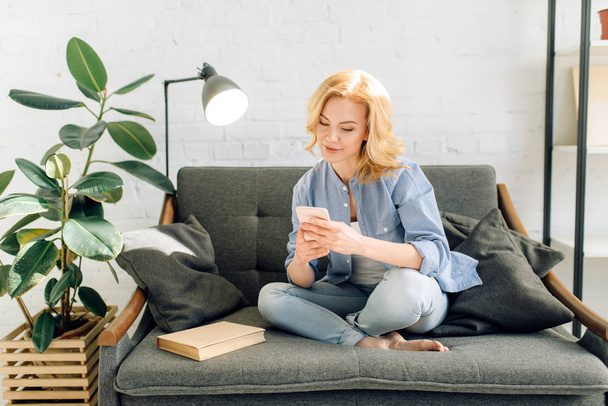 Mladá žena, která používá mobilní telefon na útulném černém gauči, obývací pokoj s bílými tóny a hrnec s květinou na pozadí. Atraktivní ženská osoba sedící na pohovce doma - Fotografie, Obrázek