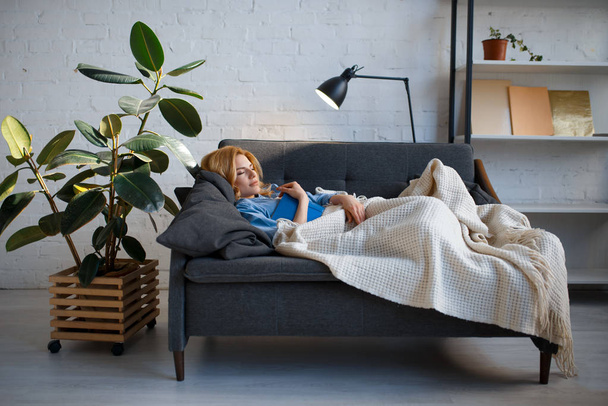 Young woman lying on cozy black couch and reading a book, living room in white tones on background. Attractive female person with magazine rest on sofa at home - Foto, immagini