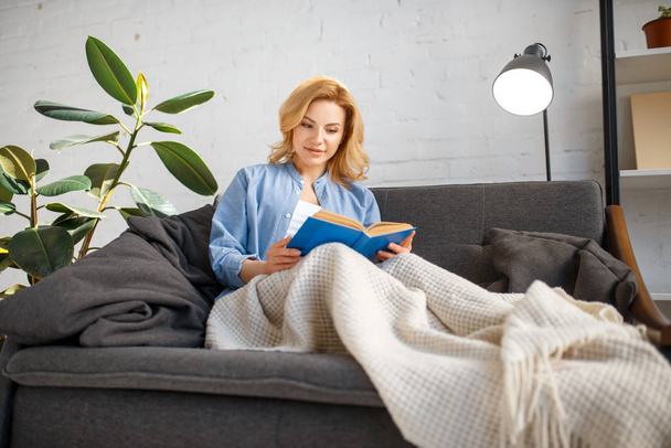Young woman under a blanket reading a book on cozy yellow couch, living room in white tones on background. Attractive female person with magazine sitting on sofa at home - 写真・画像
