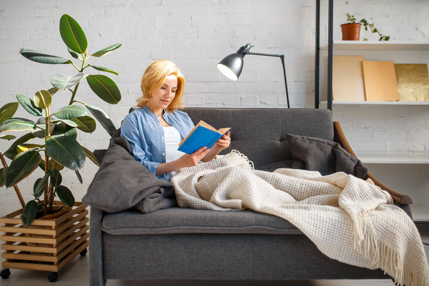 Young woman under a blanket reading a book on cozy yellow couch, living room in white tones on background. Attractive female person with magazine sitting on sofa at home - Фото, изображение