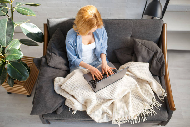 Rahat sarı kanepede laptop kullanarak bir battaniye altında genç kadın, arka planda beyaz tonlarda oturma odası. Evde kanepede oturan dergi ile çekici kadın kişi - Fotoğraf, Görsel