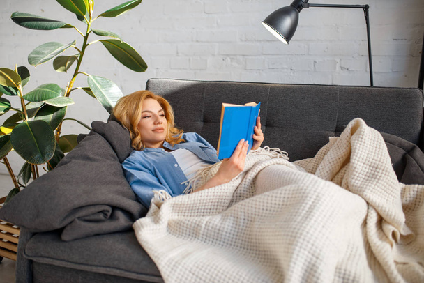Young woman under a blanket reading a book on cozy yellow couch, living room in white tones on background. Attractive female person with magazine sitting on sofa - 写真・画像