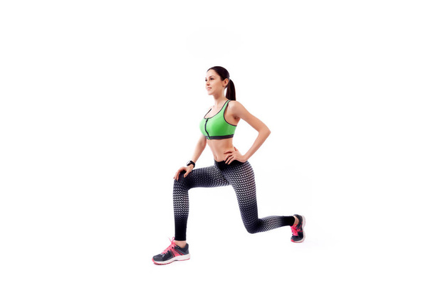 Een jonge vrouw model in een sportieve korte top en Gym legging maakt longen door de voeten naar voren, handen worden gehouden aan de zijkant op een witte geïsoleerde achtergrond in de Studio  - Foto, afbeelding