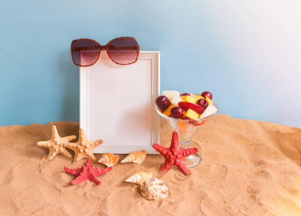 Empty frame for your text, bowl of fruit salad, sunglasses, starfishes and seashells on sand beach on blue backdrop. - Photo, Image