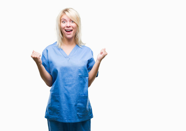 Young beautiful blonde nurse doctor woman over isolated background celebrating surprised and amazed for success with arms raised and open eyes. Winner concept. - Photo, Image