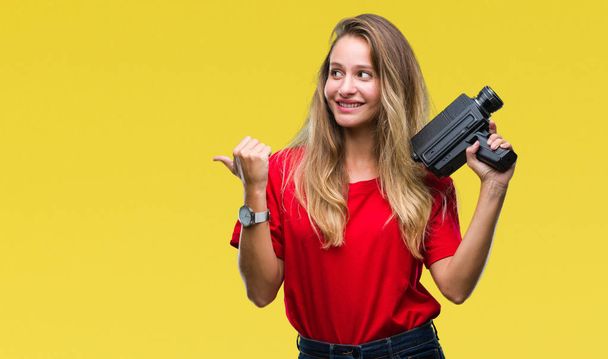 Young beautiful blonde woman filming using vintage camera over isolated background pointing and showing with thumb up to the side with happy face smiling - Foto, imagen