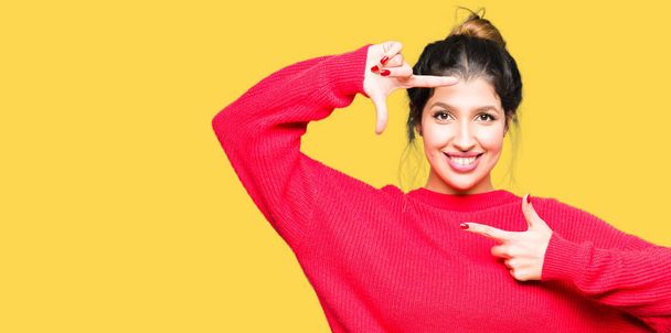 若い美しい女性の赤いセーターとお団子を手で作るフレームと幸せそうな顔で指を笑顔を身に着けています。創造性と写真のコンセプト. - 写真・画像