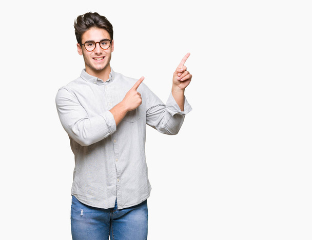 Homem bonito jovem usando óculos sobre fundo isolado sorrindo e olhando para a câmera apontando com duas mãos e dedos para o lado
. - Foto, Imagem