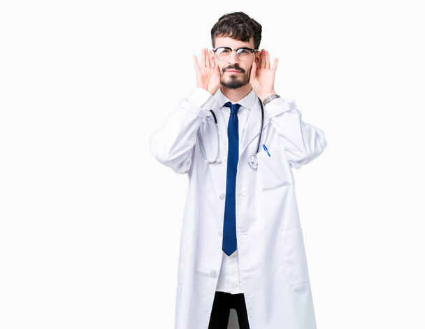 Молодий лікар в лікарняному пальто над ізольованим тлом Намагаючись почути обидві руки на жесті вуха, цікаво для пліток. Проблема зі слухом, глуха
 - Фото, зображення