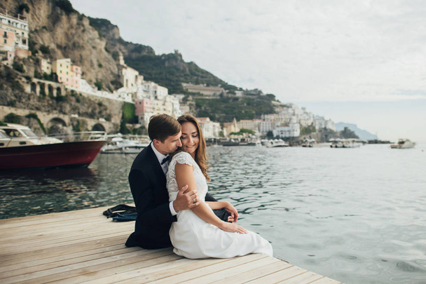 Happy romantic couple sitting on wooden pier on sea bay in Italy - Photo, Image