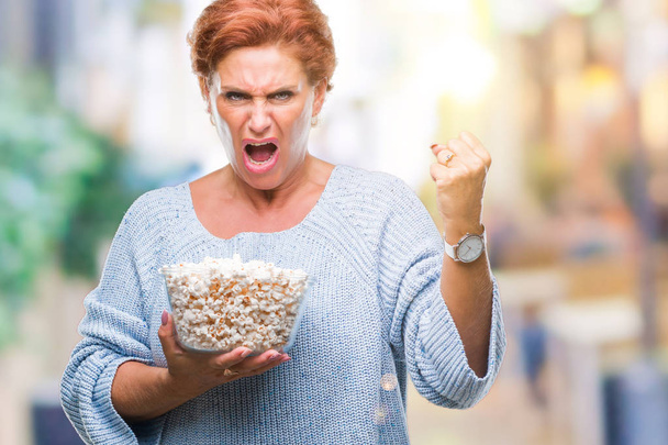 attraktive ältere kaukasische rothaarige Frau, die Popcorn vor isoliertem Hintergrund isst, genervt und frustriert schreit vor Wut, verrückt und schreit mit erhobener Hand, Wut-Konzept - Foto, Bild