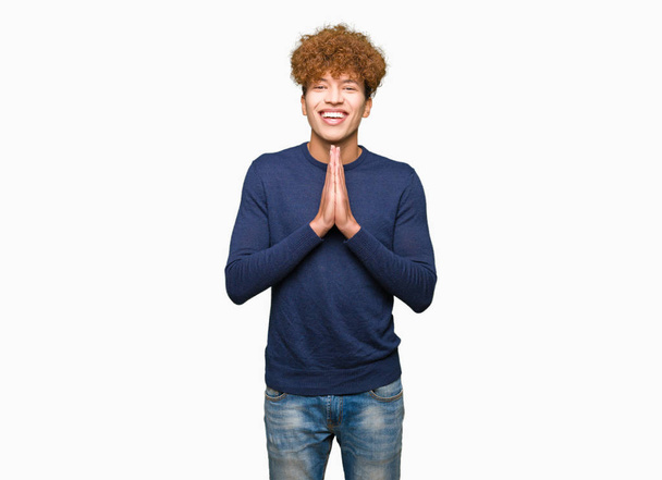 Молодой красивый мужчина с афроволосами молится руками вместе, прося прощения, уверенно улыбаясь.
. - Фото, изображение