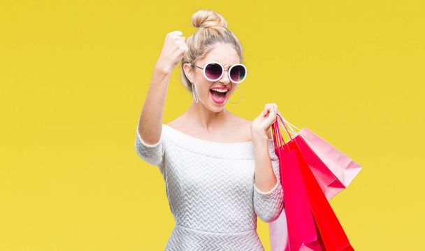 Young beautiful blonde woman holding shopping bags over isolated background annoyed and frustrated shouting with anger, crazy and yelling with raised hand, anger concept - Fotografie, Obrázek