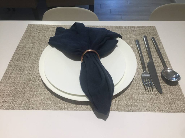 White dish on the white table with cutlery, napkin in a ring. Empty plate on a table mat with spoon fork and knife. - Foto, Bild