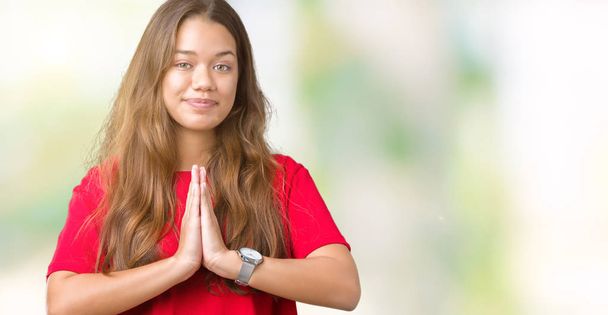 Young beautiful brunette woman wearing red t-shirt over isolated background praying with hands together asking for forgiveness smiling confident. - Photo, image