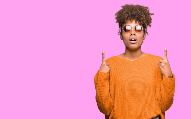 Beautiful young african american woman wearing sunglasses over isolated background amazed and surprised looking up and pointing with fingers and raised arms. - Foto, Imagem