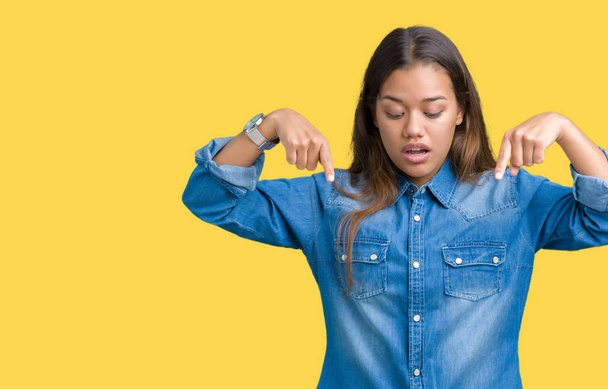 Молодая красивая брюнетка в синей джинсовой рубашке на изолированном фоне, указывающая пальцами на рекламу, удивленное лицо и открытый рот
 - Фото, изображение