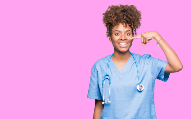 分離以上の若いアフリカ系アメリカ人医師女性は陽気な笑みを浮かべて顔や鼻、手指でポインティングを背景します。 - 写真・画像