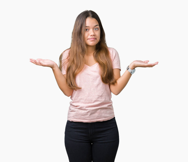 Young beautiful brunette woman wearing pink t-shirt over isolated background clueless and confused expression with arms and hands raised. Doubt concept. - Фото, зображення