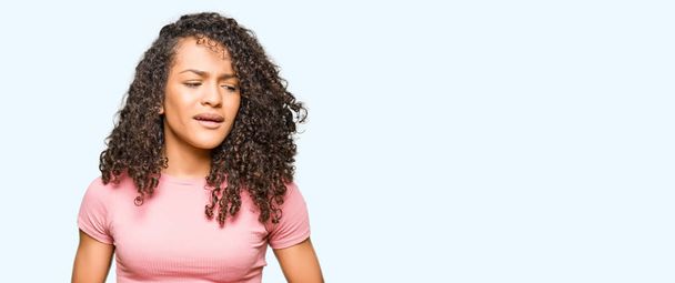 Young beautiful woman with curly hair wearing pink t-shirt with hand on stomach because nausea, painful disease feeling unwell. Ache concept. - Фото, изображение