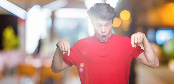 Jóképű fiatalember visel piros póló, felső elszigetelt háttér mutatva le ujjait bemutató reklám, meglepett arcát és nyitott szájjal - Fotó, kép