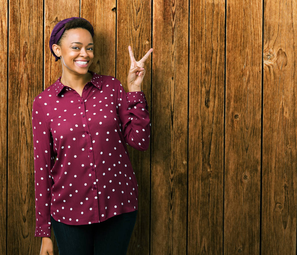 Красива молода жінка афро-американських носити шарф начальника над ізольовані фон посміхається з щасливим обличчям, підморгування на камеру, що робити знак перемоги. Номер два. - Фото, зображення