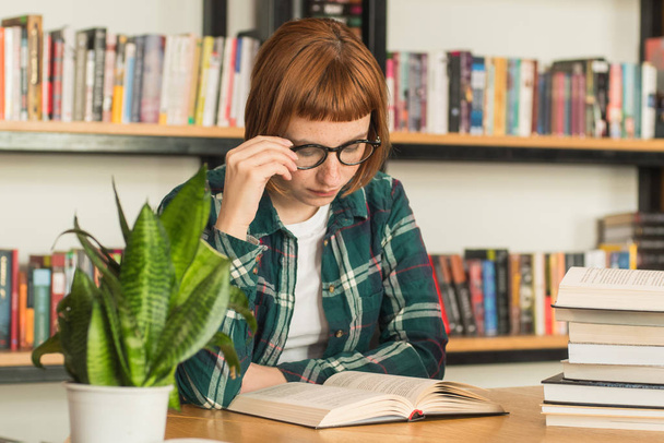 Giovane rossa donna in occhiali letto libro in biblioteca
 - Foto, immagini