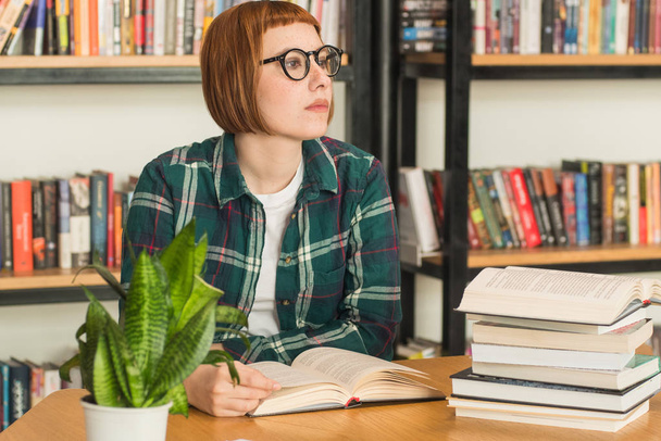 junge rothaarige Frau mit Brille liest Buch in der Bibliothek - Foto, Bild