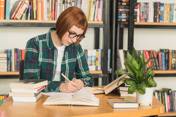 Joven pelirroja en gafas leer libro en la biblioteca
 - Foto, imagen