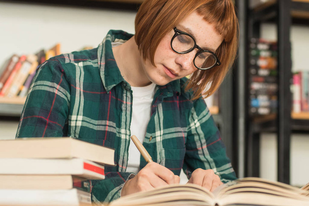 Young redhead woman in glasses read book in the library - Photo, Image