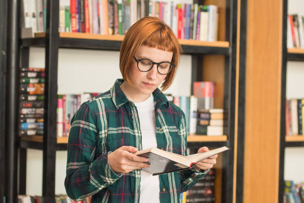 Fiatal vöröshajú nő a szemüveg könyvet olvasni a könyvtárban - Fotó, kép