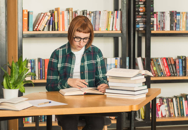 Young redhead woman in glasses read book in the library - Photo, Image