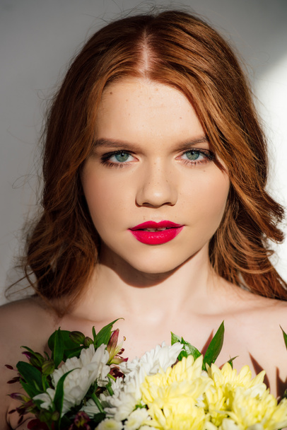 beautiful young redhead woman with red lips posing with flowers and looking at camera  - Zdjęcie, obraz