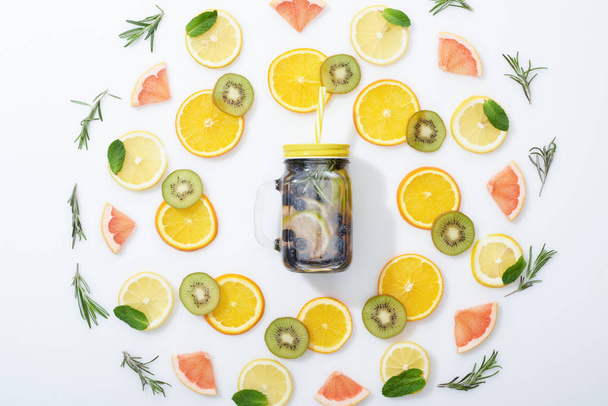 vlakke lay met gesneden kiwi, sinaasappelen, citroenen, grapefruits, munt, rozemarijn en detox drank in kruik op grijze achtergrond - Foto, afbeelding
