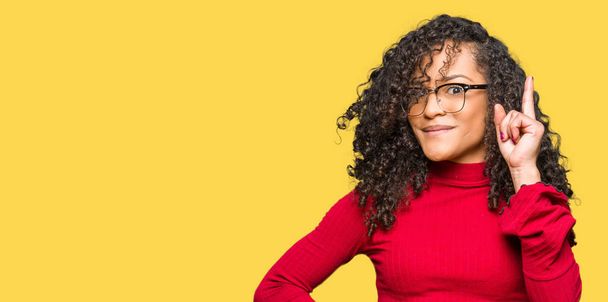 Young beautiful woman with curly hair wearing glasses pointing finger up with successful idea. Exited and happy. Number one. - Photo, Image