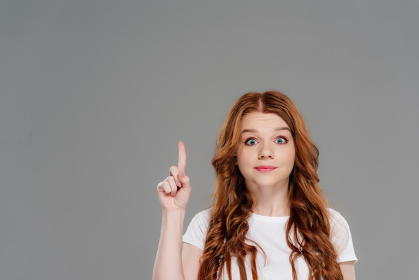 beautiful surprised redhead girl looking at camera and showing idea gesture isolated on grey with copy space - Foto, imagen